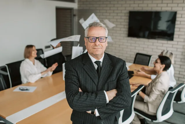 Business executive standing confidently in meeting room with team engaged in discussion behind. Ledelse i små virksomheder: ansvar, kommunikation og udvikling